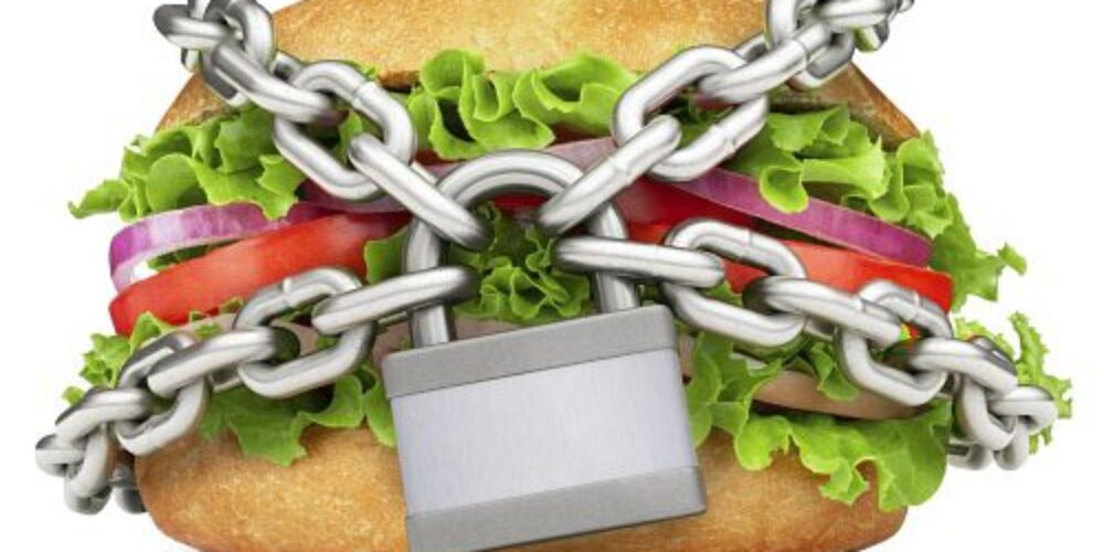 En smörgås med sallad, tomat och rödlök är inlindad i korslagda metallkedjor och låst med ett hänglås framtill, mot vit bakgrund, vilket signalerar begränsad eller förbjuden mat.